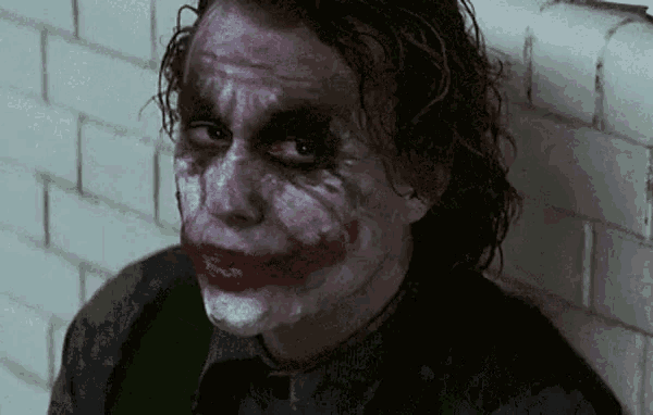 the joker is leaning against a wall with his face painted and making a funny face .