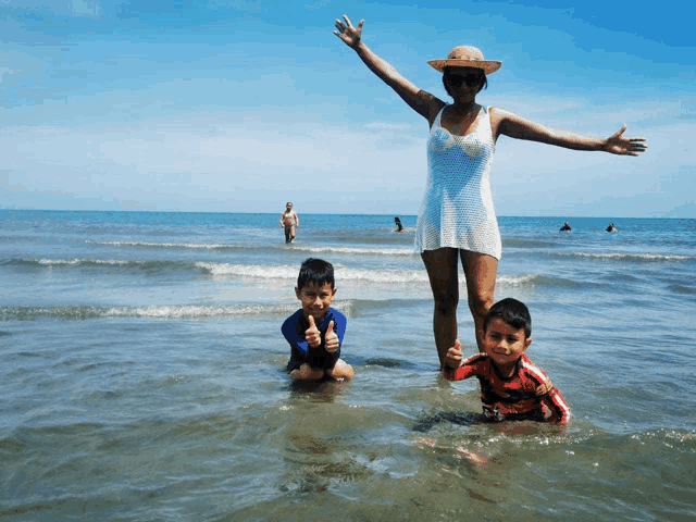 a woman in a white dress and straw hat stands in the ocean with two children
