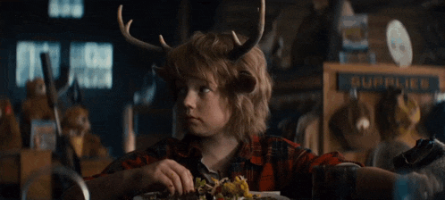 a little boy with antlers on his head sits at a table with a plate of food in front of a sign that says supplies