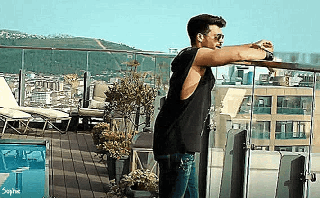 a man in a black tank top and sunglasses is leaning on a railing on a balcony overlooking a city .