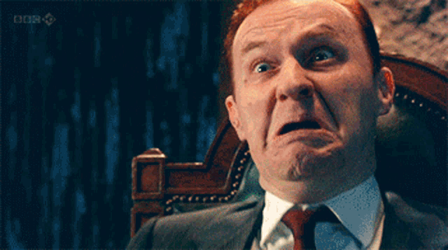 a man in a suit and tie is sitting in a chair and making a funny face with the bbc logo in the background