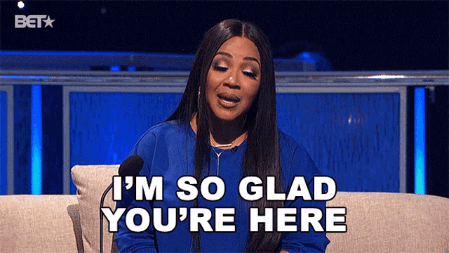 a woman in a blue shirt is sitting on a couch and says i 'm so glad you 're here .