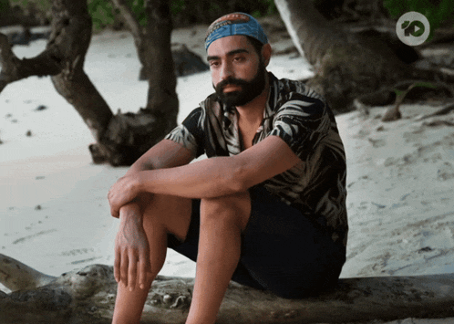 a man with a beard sits on a log on the beach with an io logo behind him