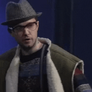 a man wearing a hat and glasses is standing in front of a blue curtain and making a funny face .