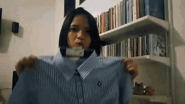 a woman is holding a blue striped shirt in front of a bookshelf filled with books .
