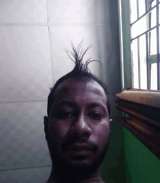 a man with a mohawk looks at the camera with a green wall in the background
