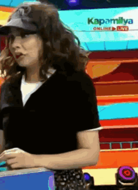 a woman wearing a black shirt and a hat is standing in front of a sign that says kapamilya online live