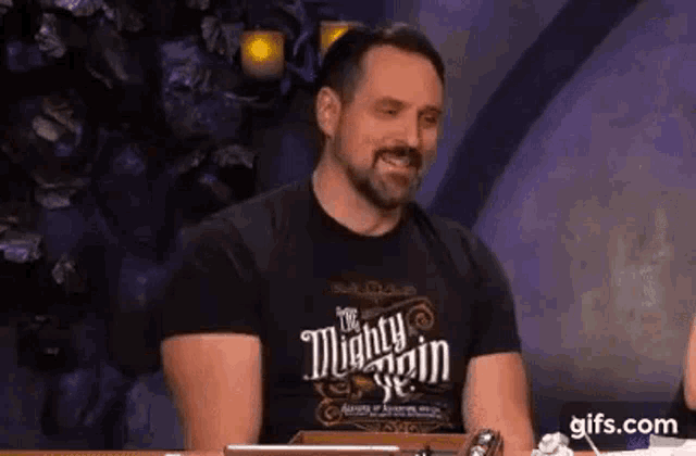 a man with a beard is sitting at a table wearing a mighty main t-shirt and smiling .