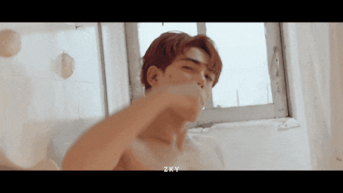 a shirtless man brushes his teeth in front of a window with zky written on the bottom right