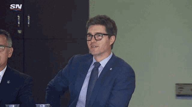 a man in a suit and tie sits at a table with a sn logo in the background