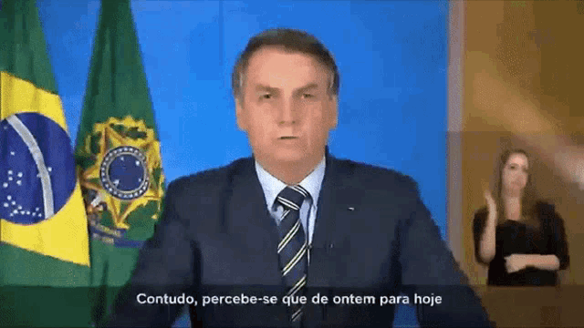 a man in a suit and tie is giving a speech in front of two flags and says " contudo "