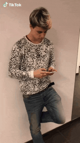 a young man leaning against a wall looking at his phone with a tiktok watermark on the bottom right