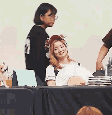 a woman is sitting at a table holding a fan while a man stands behind her .