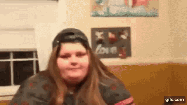 a woman wearing a baseball cap is making a funny face while sitting in front of a window .