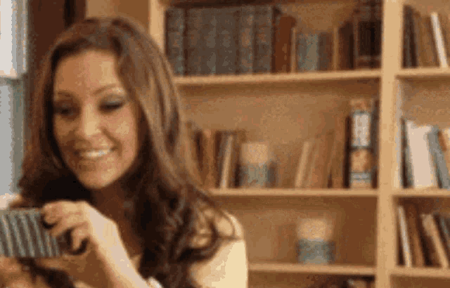 a woman is smiling while holding a cell phone in front of a bookshelf