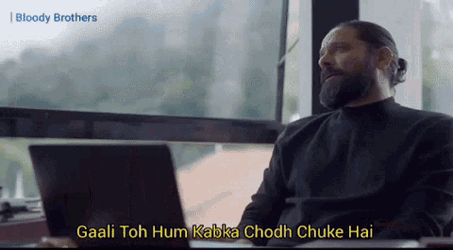 a man with a beard is sitting in front of a laptop computer and says " gaali toh hum kabka chodh chuke hai "