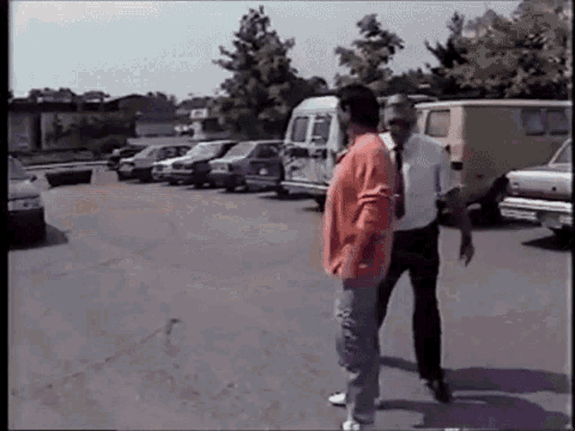 two men are standing next to each other in a parking lot with cars parked in the background .