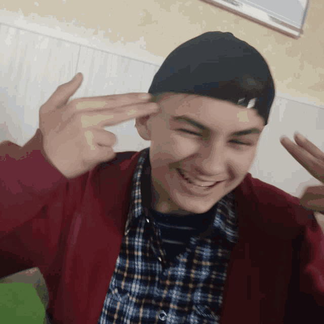 a young man wearing a plaid shirt and a red jacket is smiling and pointing at his head