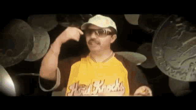 a man wearing sunglasses and a yellow shirt with the word hard knocks on it is pointing at his head .