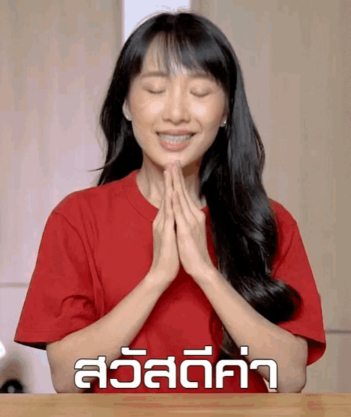 a woman in a red t-shirt prays with her eyes closed in front of a sign that says สวัสดี ค่า