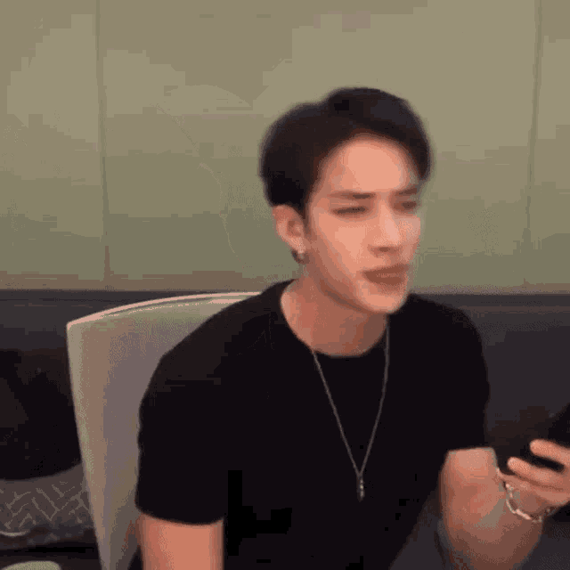 a young man wearing a black shirt and a necklace is sitting in a chair holding a cell phone .