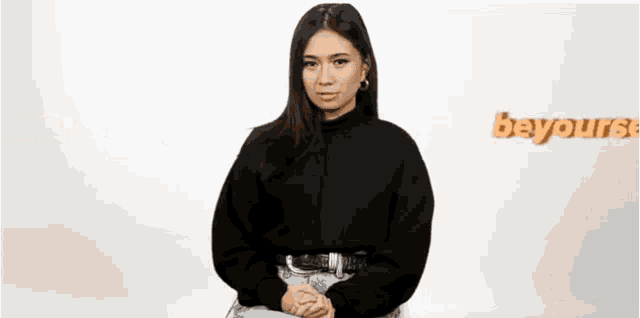 a woman in a black sweater and jeans is standing with her hands folded in front of a white wall .