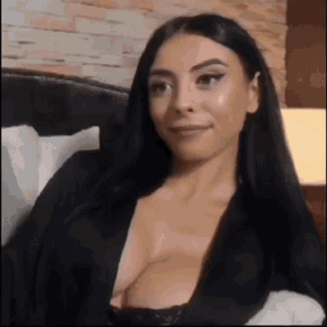 a woman with long black hair is sitting on a bed with her breasts showing .