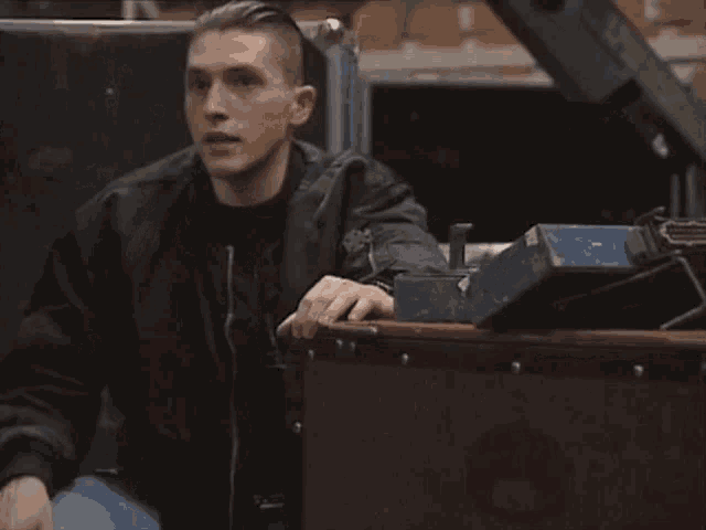 a man in a black jacket is sitting in front of a wooden box that says ' a ' on it