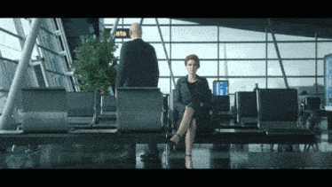 a woman sits in an airport waiting area while a man stands in front of a sign that says gate