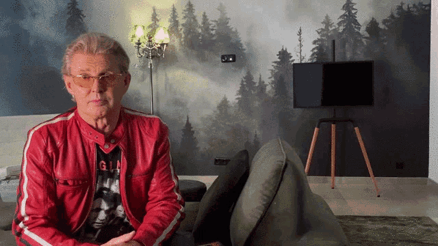 a man in a red jacket is sitting on a couch in front of a wall that has trees on it