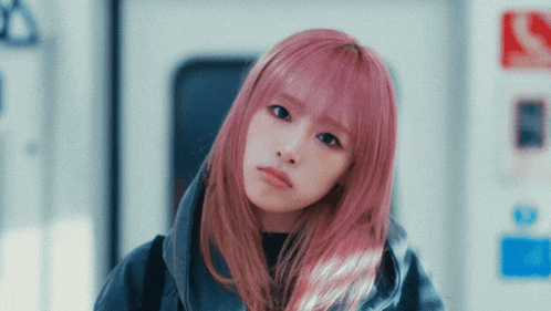 a girl with pink hair looks at the camera with a sign in the background that says in
