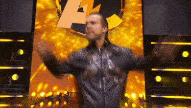 a man in a leather jacket is standing on a stage with his arms outstretched in front of a yellow background .