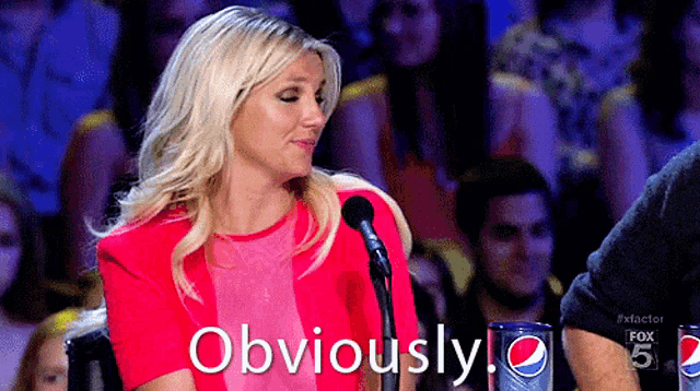 a woman in a pink shirt is sitting in front of a microphone and the word obviously is on the screen behind her