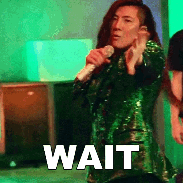a woman in a green sequined jacket is singing into a microphone and the word wait is on the screen behind her .
