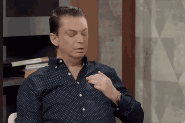 a man in a blue shirt is sitting in a chair with his eyes closed and his hand on his chest .