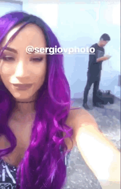 a woman with purple hair is taking a selfie with a man standing behind her looking at his phone