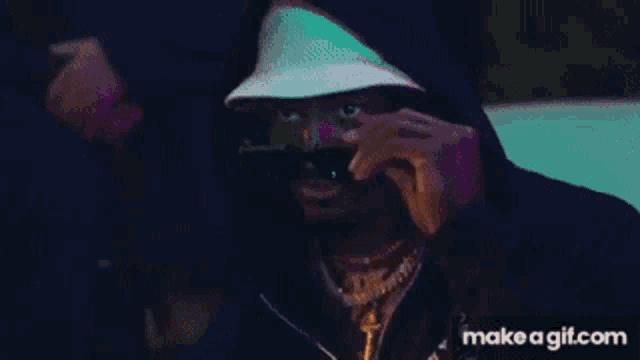 a man wearing a hat and sunglasses is covering his face with his hand .
