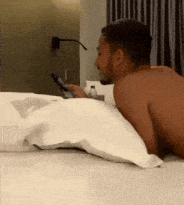 a shirtless man is laying on a bed looking at a phone