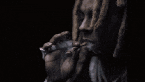 a man with dreadlocks is smoking a cigarette in a dark room with smoke coming out of his mouth .