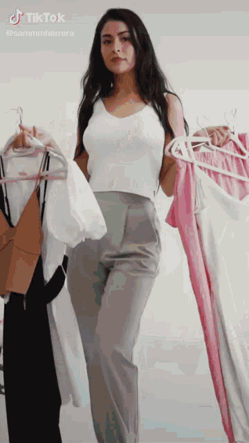 a woman in a white tank top and grey pants is holding clothes in front of a white wall with a tiktok logo above her