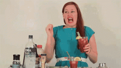 a woman is sitting at a table holding a bloody mary and making a face .
