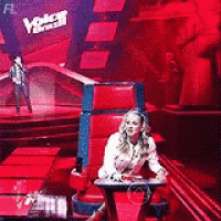 a woman is sitting in a red chair on a stage with a man standing in the background .
