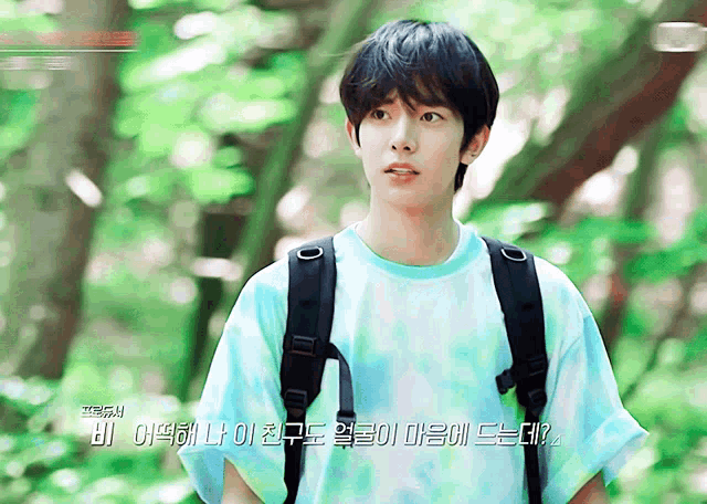 a young man wearing a blue tie dye shirt and a black backpack stands in a forest