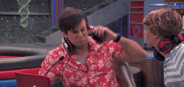 a man in a hawaiian shirt is sitting in front of a microphone while another man looks on .