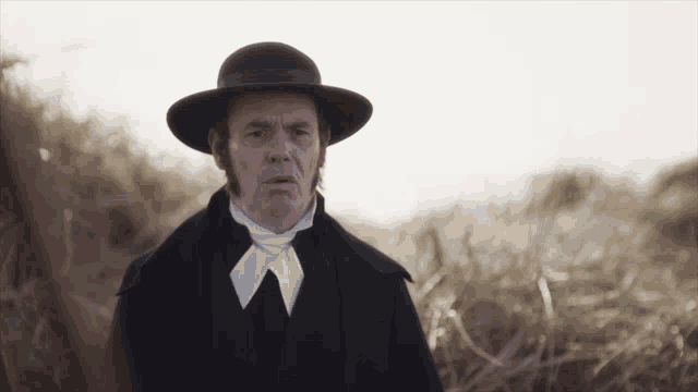 a man wearing a black hat and a black coat stands in a field