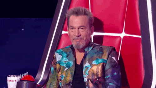a man with a beard is sitting in a chair with a red curtain behind him .