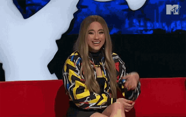 a woman in a colorful jacket is sitting on a red couch with a mtv logo in the background