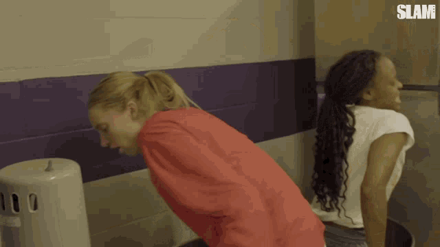 a woman in a red shirt sits next to another woman in a white shirt in a bathroom with the word slam on the wall