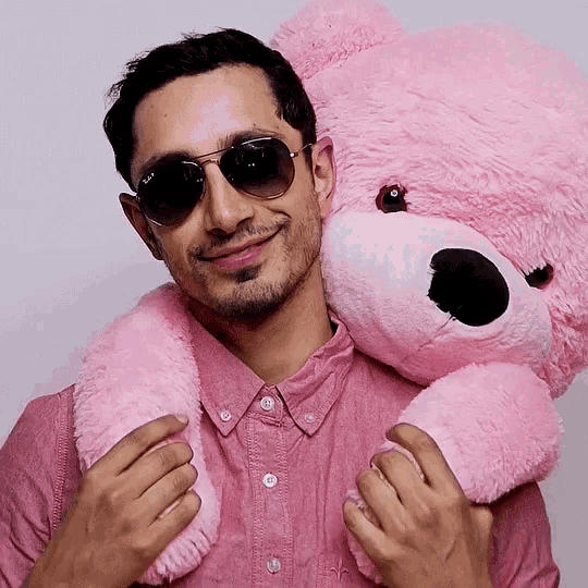 a man wearing sunglasses and a pink shirt holds a pink teddy bear