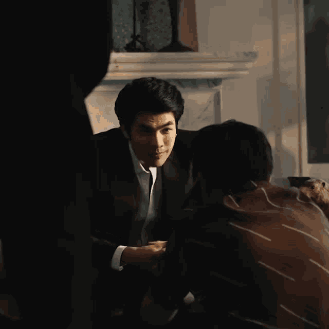 a man in a suit is talking to another man in a dark room